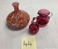 Photo showing all three items: orange and white swirled vase, plum-colored perfume bottle with stopper, and matching pitcher.