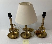 Full view of three brass lighting pieces: one lamp with beige shade and two candle holders.