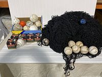 Baseball netting with multiple used baseballs scattered on it and a small blue ball caught in the netting. Several new boxed Worth baseballs are also visible.