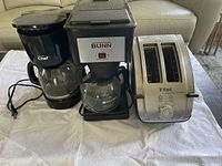 Photo showing three kitchen appliances on a white towel: a Bunn commercial coffee maker, a black Chef drip coffee maker, and a stainless steel T-fal toaster.