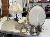 Photo showing vintage-style table lamp with cream fabric shade and fringes, two ceiling lamps with frosted glass shades, and new boxed ceiling light fixture components on table.