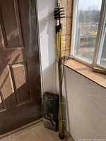 Eight yellow 5-foot rods leaning against wall with several black chimney brushes attached and a black metal carrying case on floor.
