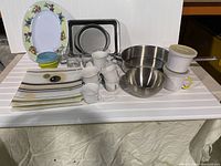 Overview of frying pans, baking sheets, ceramic plates, coffee cups, stainless steel mixing bowl and stacking ceramic pots.