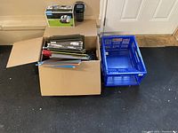 Open cardboard box with assorted coloured hanging file folders and a blue collapsible file holder placed beside it on the floor, handheld label maker and boxed Dymo LabelWriter 450 visible on top