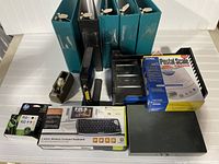 Photo of various office supplies on white background including binders, postal scale, keyboard, ink cartridges, cash box and other desk accessories.