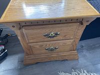 Side view of an oak finish night stand showing carved detailing, two drawers with ornate brass-colored handles, and overall condition.