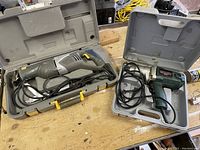 Photo of two corded electric power tools each in their own plastic carrying case on a workbench. Left is a reciprocating saw with yellow accents, right is an impact tool in dark green color.