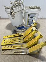 Four yellow metal roof brackets lined up, two buckets containing ropes with metal safety latches, and multiple metal hardware components on floor