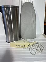 Photo showing stainless steel waste container, ironing board, and Eureka handheld vacuum cleaner with cord on a white surface.