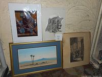 Four vintage art pieces positioned on floor: colorful African print with figure and jewelry, pencil Egyptian stone carving profile, desert landscape with palm trees in frame, and 1890s English engraving mounted on mat.