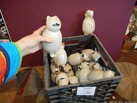 Photo showing a hand holding a ceramic cat-shaped pitcher or vase, cream color, and many similar cat figurines in a crate behind. Shows detailed shape and size of individual cat-shaped ceramics.