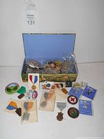 Photo of open box with various pins, medals, and badges displayed inside and around it, showing colorful ribbons and enamel badges, some individually bagged.