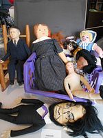 Wide view of the boxlot showing the 1960s JFK vinyl doll sitting on wooden chair among multiple handmade dolls in cloth, porcelain, and crochet.