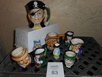 Image of nine miniature Toby jugs and mugs on a black lattice surface with a white auction card labeled 83. The jugs depict various characters including a pirate with an eye patch and skull hat, sailors with white hats, and several other colorful faces.