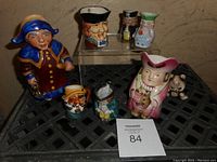 Overview photo showing eight mini Toby jugs and pitchers arranged on a surface, highlighting various characters, colors, and sizes for the lot.