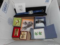 Box with multiple costume pieces including watches, carved jade earrings, Bob Hope caricature lighter and keyring, gold-tone earrings and chain.