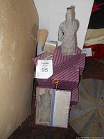 Single terracotta warrior figurine displayed on top of a decorative purple-patterned gift box. Another identical figurine is partially visible inside its gift box with the lid open.