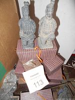 Two kneeling terracotta warrior figurines sitting on patterned gift boxes inside a cardboard box, some dust and wear visible on figurines and boxes.