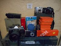 Lot showing Canon AE-1 film camera, two digital cameras, IBM tape box, orange storage containers, appliance timer, camera flash, Zikk timer, and Canon box