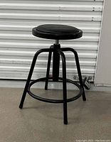 Front angle view of black adjustable stool showing padded seat and metal legs with footrest ring.