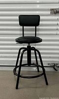 Front view of the black adjustable stool with padded round seat and backrest against a white corrugated background.
