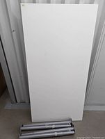 White rectangular desktop with edges visible showing wear, paired with three silver metal legs stacked beside it on floor.