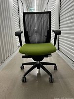 Front view of black and green office chair with mesh back and green cushioned seat, showing armrests and caster wheels.
