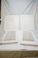 Four white wooden window shutters positioned on a white cloth background. Two shutters are larger and two are smaller, all with horizontal slats. Some shutters have a built-in blind mechanism behind the slats.