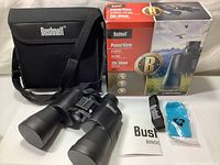 Full set shown including binoculars with lens caps, carrying case with strap, Bushnell neck strap, instruction manual, and sealed blue lens cleaning cloth in front of original packaging.