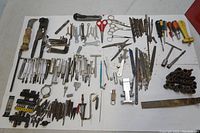 A wide assortment of various hand tools including drivers, punches, Allen keys, screwdrivers, scissors, and saw blades spread out on a white surface.