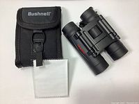 Front view of Tasco binoculars next to Bushnell carrying case and cleaning cloth on white surface.