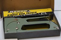 Green metal staple gun tacker in original yellow and black box showing the product in the box, highlighting the tool and packaging condition.
