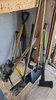 Photo showing several tools including pick axe, axes, shovel, and rake leaning against the wall.