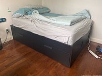 Side view of black double bed frame with built-in drawers, displaying fitted mattress sheet and bedding on top, with visible scratches on frame surface.
