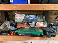 Shelf overview of closed green Coleman stove, tent bags, screen house bag, propane cylinders, pegs, inverter and cleaning tools