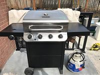 Front view of Char-Broil grill showing stainless steel hood, control panel with four knobs, black cabinet base and one propane tank