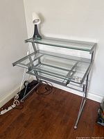 Glass top desk with metal frame, two tiers and slide-out keyboard shelf. Small plastic desk lamp on upper shelf.