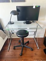 Front view of glass desk with black rolling stool, black desk lamp, Samsung monitor on desk.