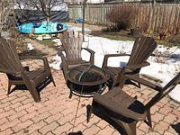 Four brown resin Muskoka chairs arranged around a small fire pit outside on a brick patio with snow in the background.