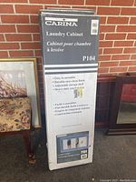 Single unopened Carina P104 laundry cabinet box leaning against brick wall showing brand and product name.