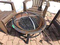 Photo showing top view of round metal outdoor fireplace with rust and mesh dome cover, alongside brown patio chairs.