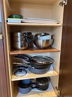 Kitchen cupboard with stacked stainless steel pots and mixing bowls on upper shelves, non-stick pans with glass lids on middle shelf, and smaller pots on bottom shelf