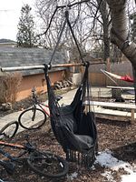 Black canvas swing hammock hanging outdoors attached to metal or wooden bars with black fringes around bottom edge shown with bicycles and yard in background