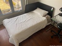 Side angle of the assembled single twin bed in a room by the window showing the headboard, mattress with white bedding, and part of the wooden floor beneath.