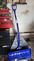 Blue plastic snow shovel with D-handle leaning against a black cabinet near a fan rake head on the floor.