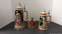 Two German beer steins and a small glass flask with leather cover displayed together on black surface with white background
