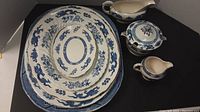 Set of large blue and white china platters stacked with gravy boat, soup tureen with lid, and small cream pitcher on black surface