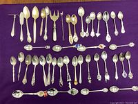 Top-down image showing approx 40 spoons and forks arranged on purple cloth, featuring a variety of styles including souvenir spoons and serving utensils.