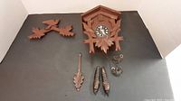 Overall view of cuckoo clock disassembled with housing, carved birds, leaves, weights, pendulum and chains laid out on dark surface