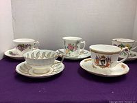 Five cups and saucers arranged showing different floral and commemorative designs.